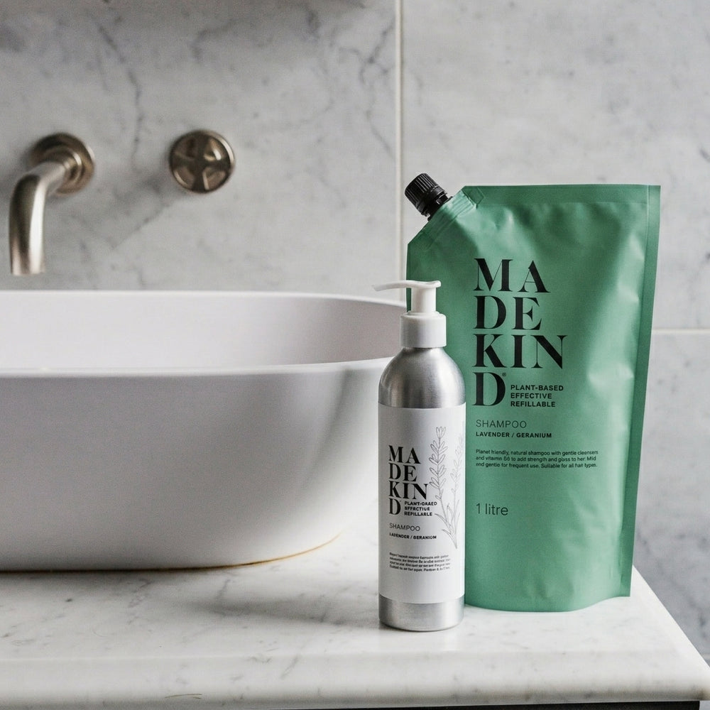 MadeKind shampoo bottle and a large green shampoo 1L pouch placed on a white marble bathroom counter next to a white vessel sink with a brass faucet.