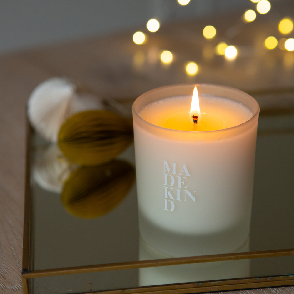 Candle with 'MADEKIND' branding on a reflective surface with blurred lights in the background