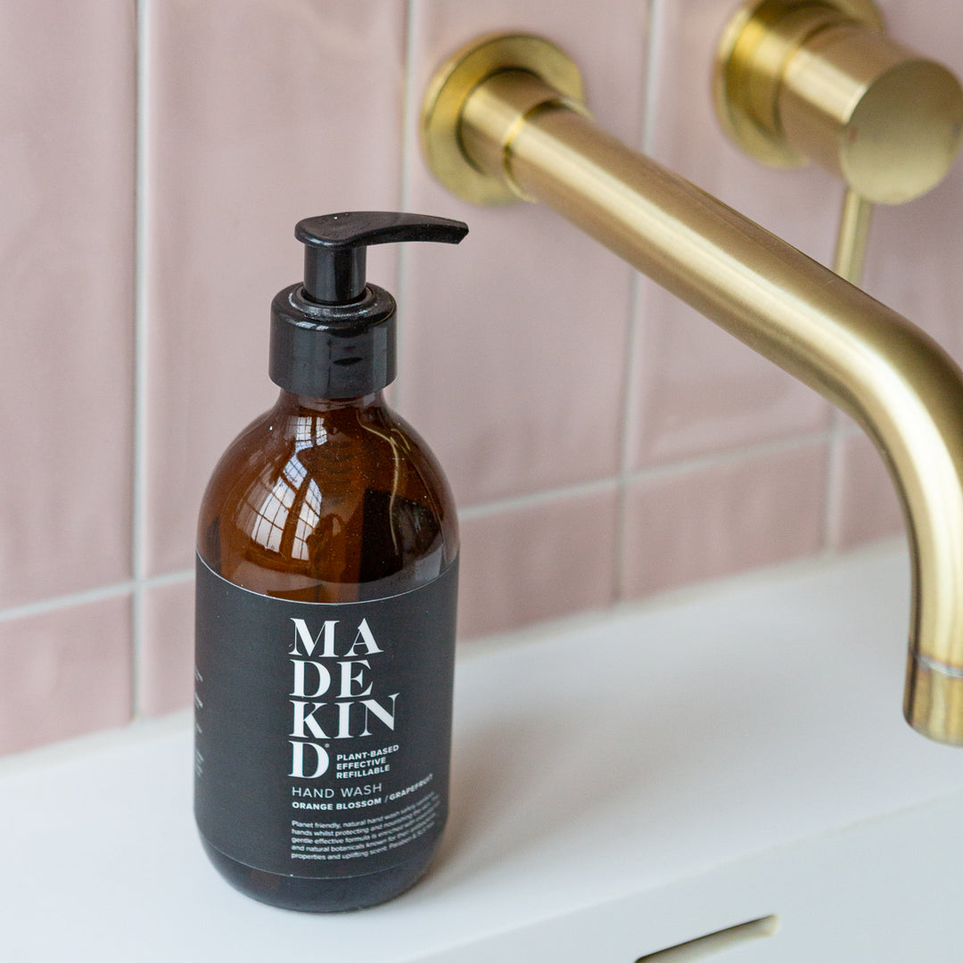 An amber glass hand wash bottle with a black pump dispenser and black label reading MadeKind, placed on a white sink counter in front of pink tiled walls and a gold tap.