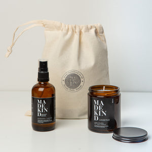 Aromatherapy amber glass unwind candle with the lid off and room mist spray in front of a madekind gift bag