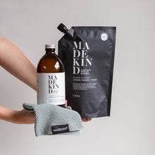 Load image into Gallery viewer, Photo of hands holding MadeKind Floor Cleaner refill pouch and an empty amber glass bottle and an organic cotton dish cloth
