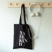Load image into Gallery viewer, MadeKind black tote bag hanging on a peg 
