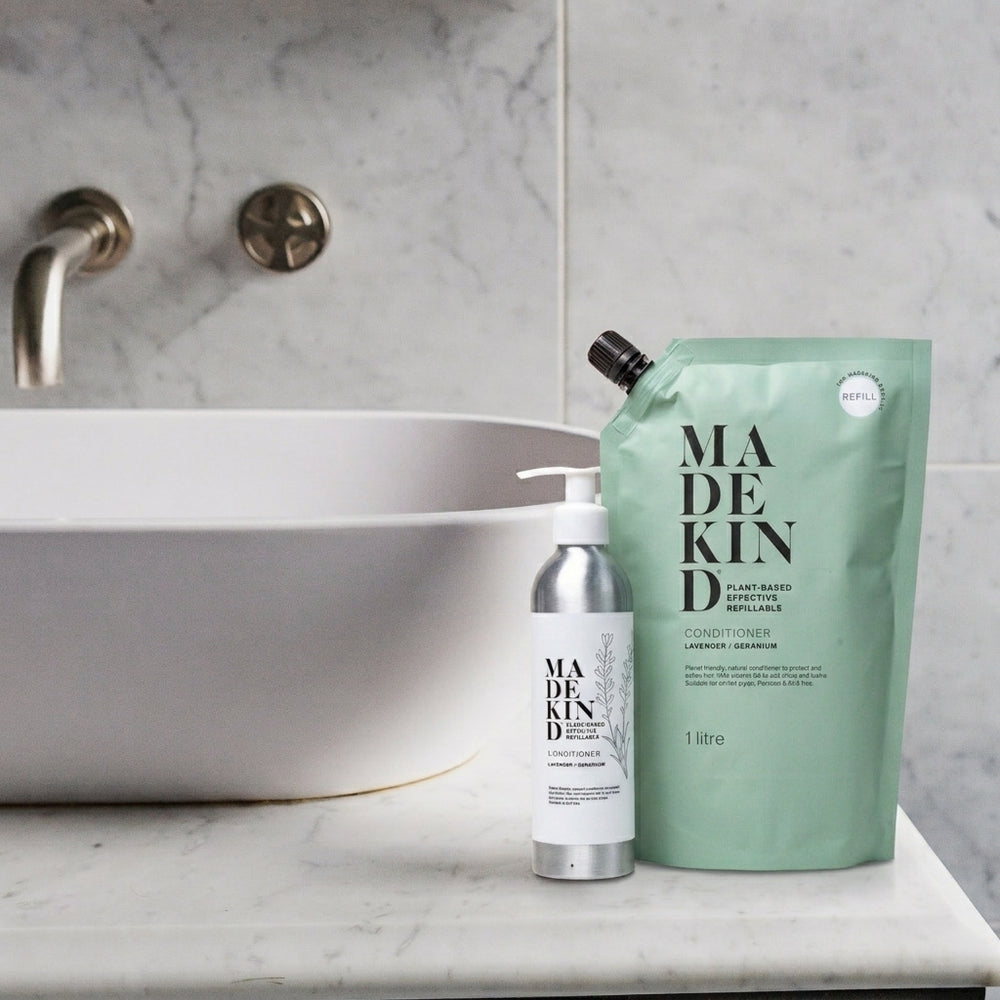 MadeKind natural haircare products shown on a bathroom counter: a silver pump bottle and a mint green 1L refill pouch beside a white sink in a marble bathroom.