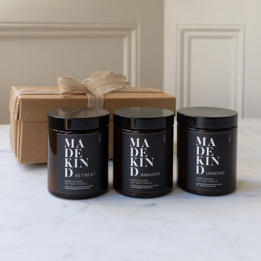 Three MadeKind soy wax candles candles in amber glass jars with black lids are arranged on a light marble surface, with one candle lit in the centre. A kraft gift box with a burlap ribbon sits behind them.