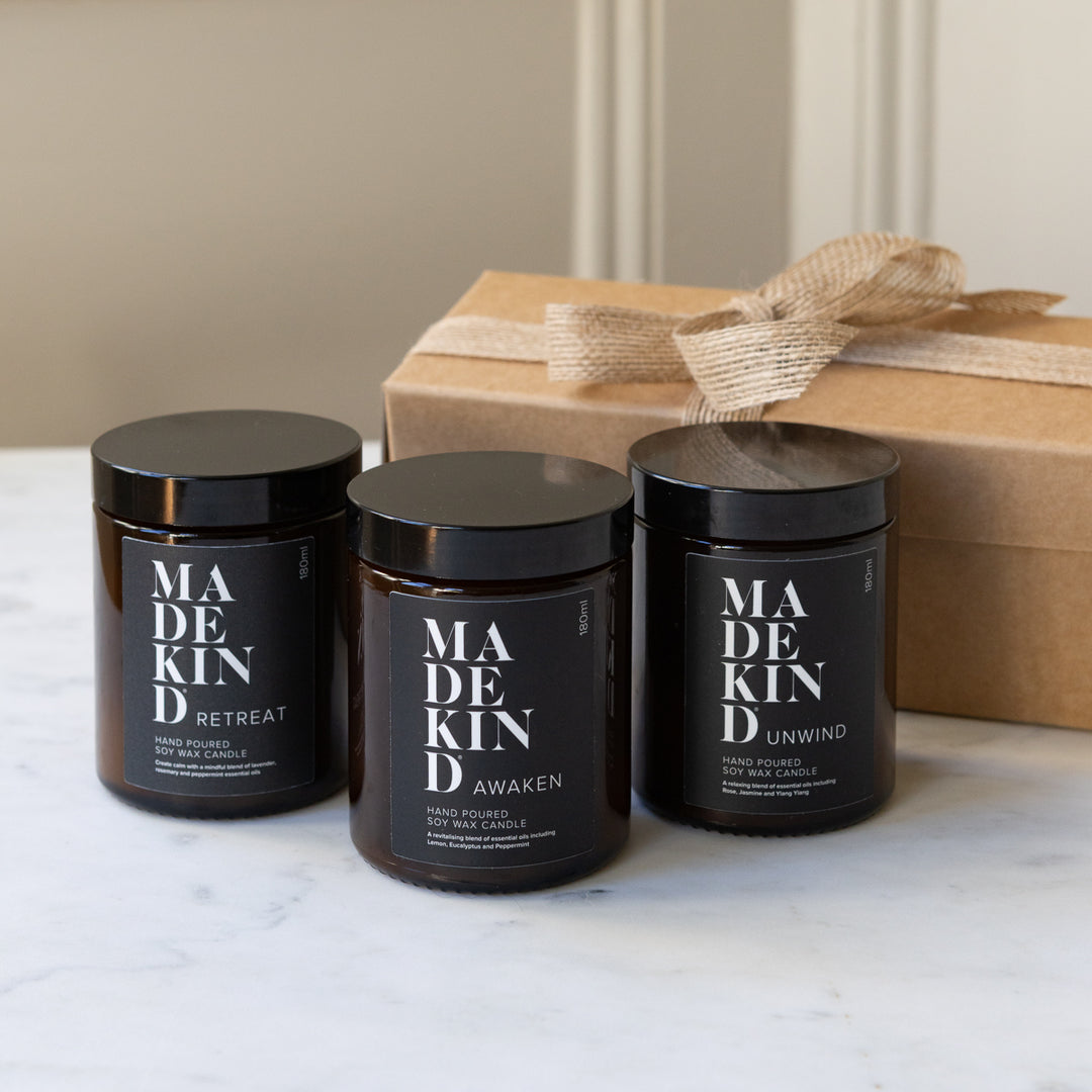 Three MadeKind soy wax candles candles in amber glass jars with black lids are arranged on a light marble surface, with one candle lit in the centre. A kraft gift box with a burlap ribbon sits behind them.