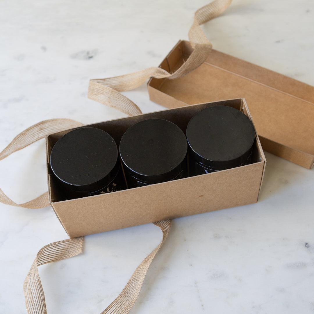 Three MadeKind soy wax candles candles in amber glass jars with black lids are arranged on a light marble surface, with one candle lit in the centre. A kraft gift box with a burlap ribbon sits behind them.