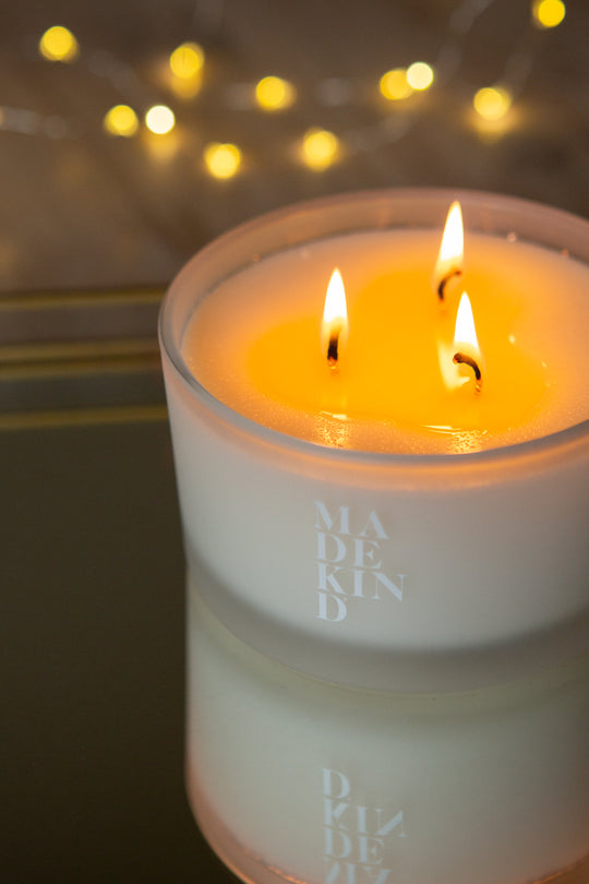 Three-wick sleep well candle in a white glass bowl with 'MadeKind' branding on the side. Lit candle with Christmas sparkly lights in the background.