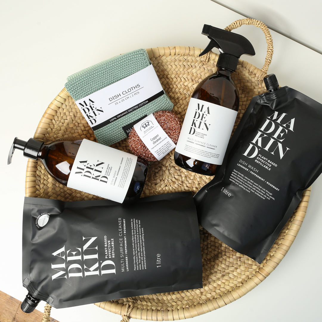 Photo of the MadeKind Kitchen Cleaning Kit in a basket; Multi Surface Cleaner and Dish Wash refills with 2 empty amber glass bottles, copper scourers and green dishcloths