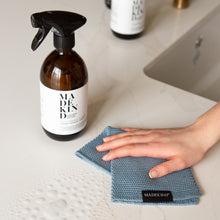 Load image into Gallery viewer, Photo of a hand holding a blue organic cotton dish cloth and a bottle of MadeKind Multi Surface Cleaner on a kitchen counter
