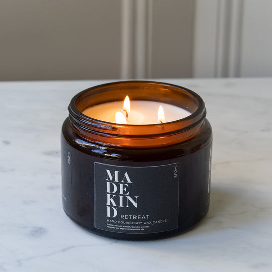 A lit amber glass jar MadeKind soy wax candle on a white marble surface with a neutral indoor background. Scented with Retreat essential oils.