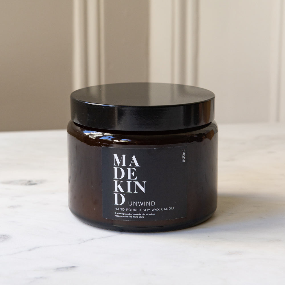 An amber glass jar MadeKind soy wax candle with three wicks on a light marble surface. The jar is amber and lid is black.
