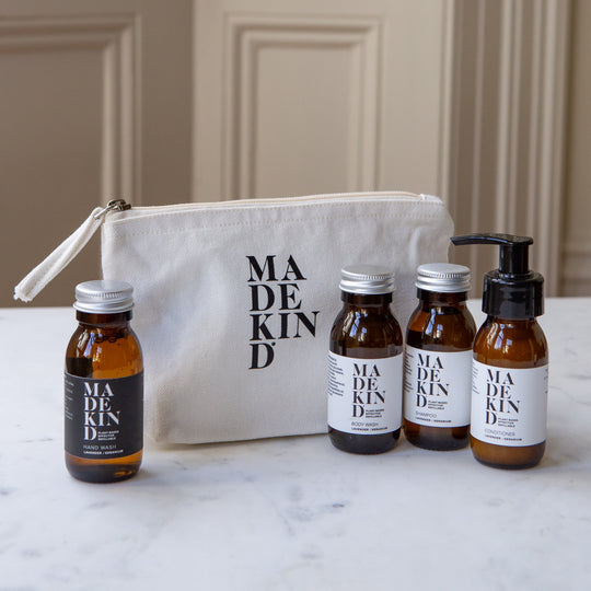 Toiletries set arranged on a white marble surface in front of a cream canvas pouch. The set includes four amber glass bottles and a pump bottle with white and black labels reading MADE KIND, plus a cream zip pouch with black MADE KIND branding.