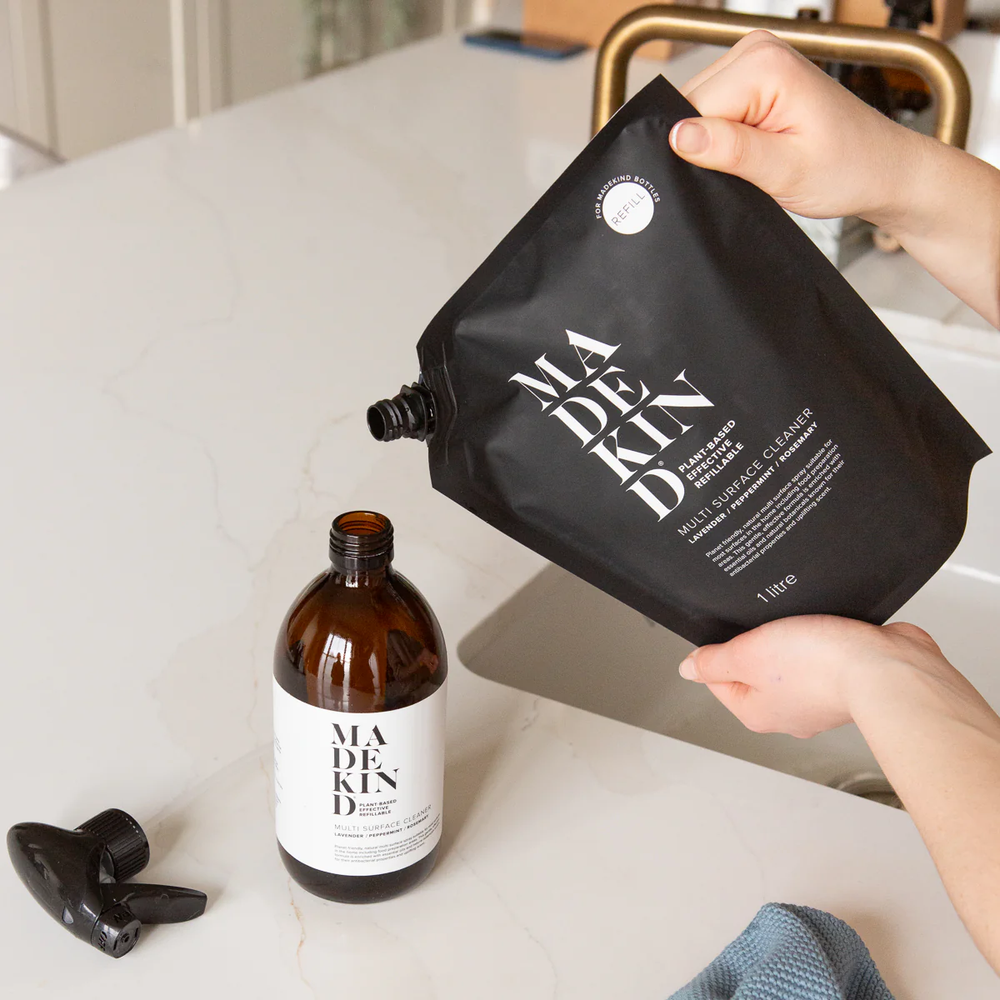 Person holding a black MadeKind refill pouch and pouring into an empty amber glass MadeKind Multi Surface cleaner bottle