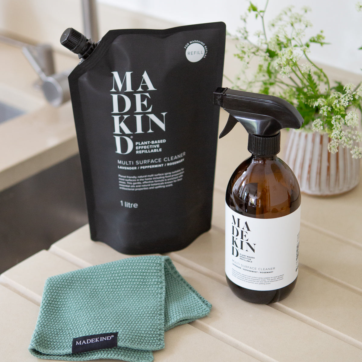 Photo of a Multi Surface Cleaner 1 Litre Refill pouch with an empty Multi Surface Cleaner bottle and green organic cotton dish cloth on a kitchen draining board.