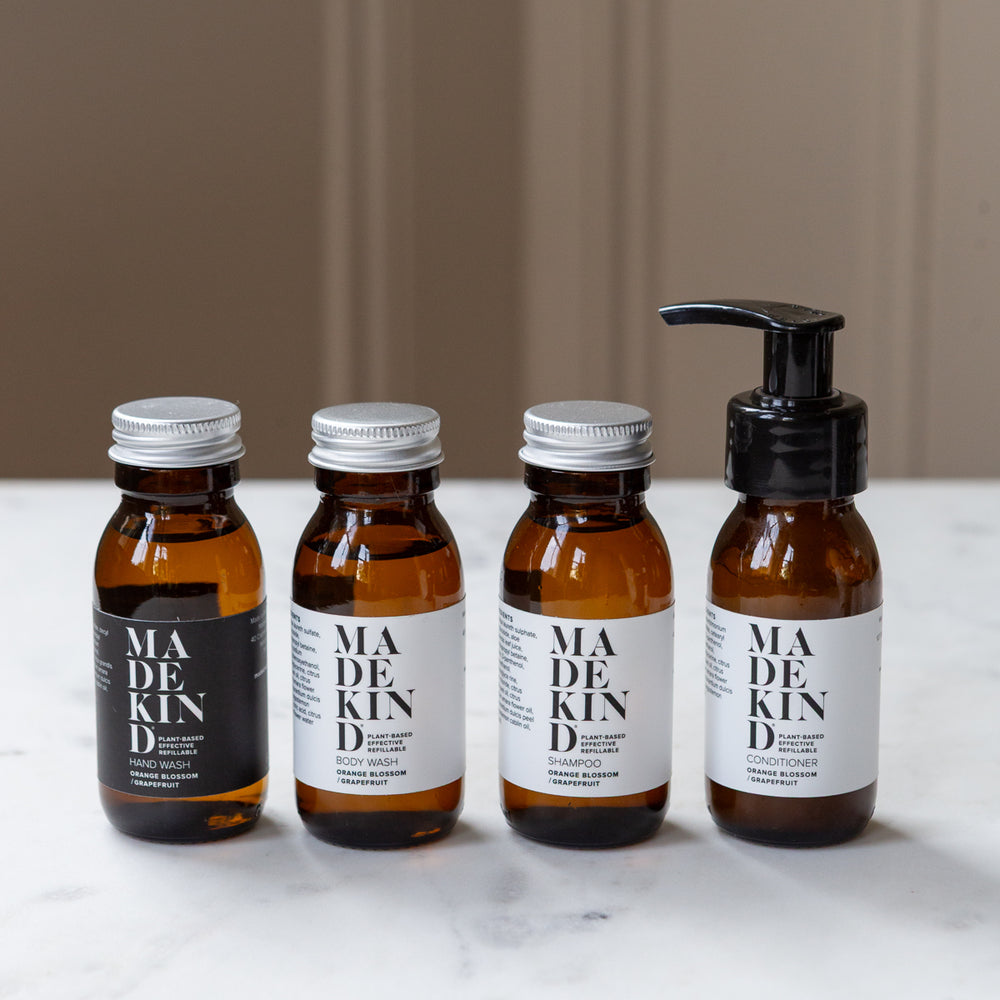 A set of MadeKind mini cleaning products arranged on a white marble surface. The set includes four amber glass bottles with cleaning products in, with white labels and metal caps, plus a cream canvas zip pouch with black text.