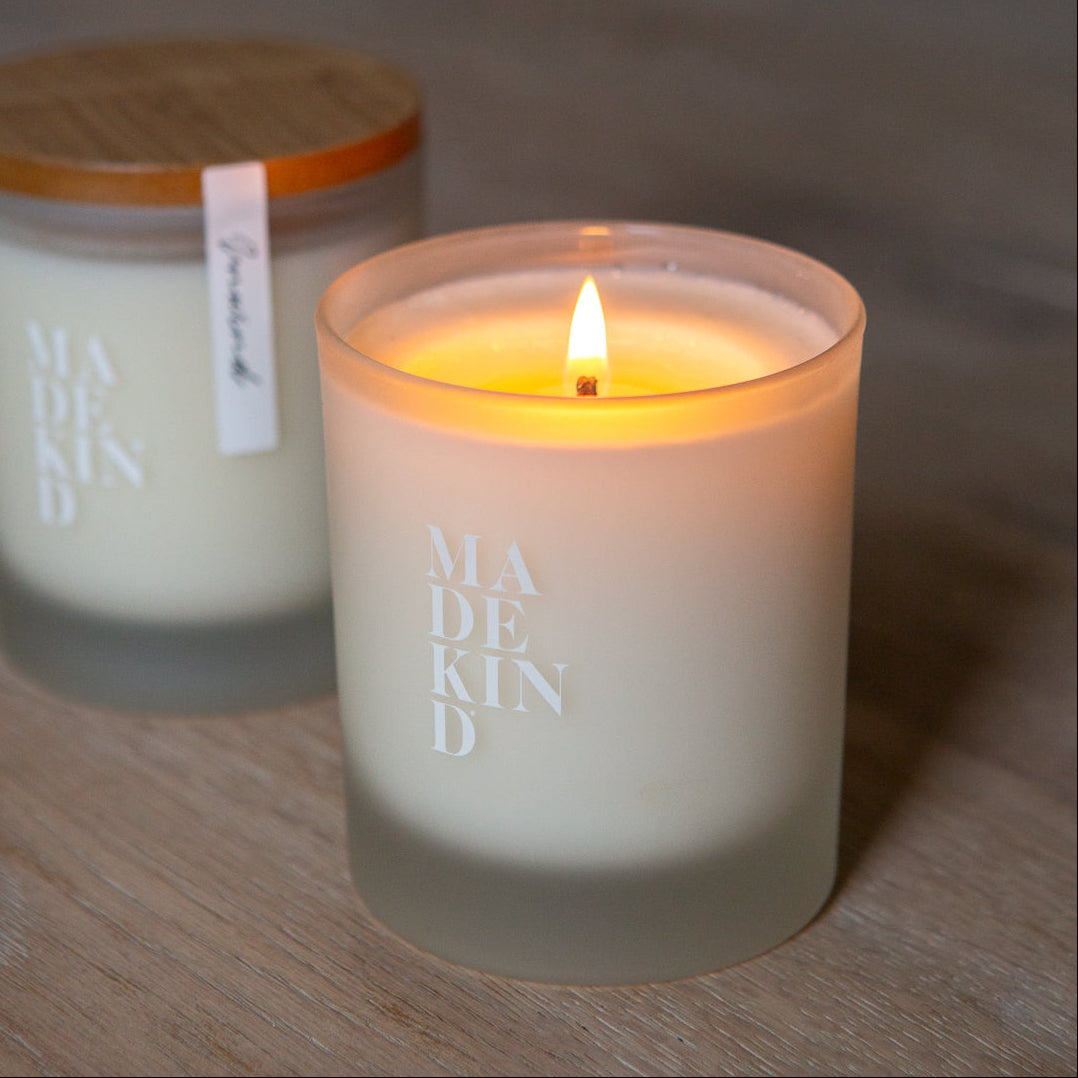 Two candles, one lit and one unlit, on a wooden surface with 'MADEKIND' branding, in unwind blend for relaxation
