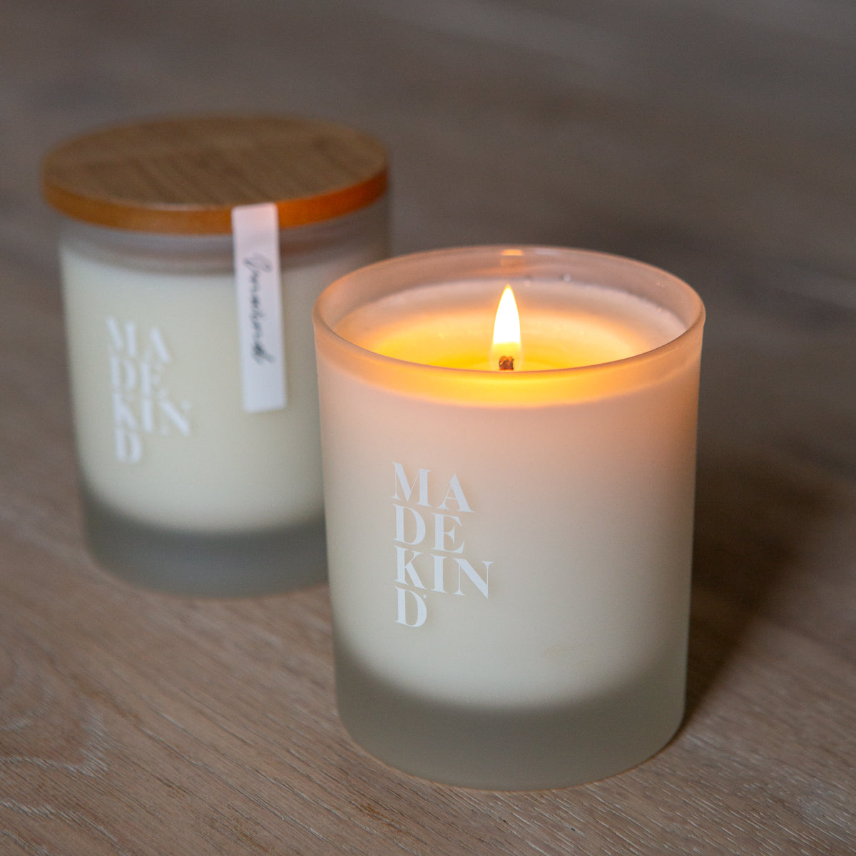 Two candles, one lit and one unlit, on a wooden surface with 'MADEKIND' branding, in unwind blend for relaxation
