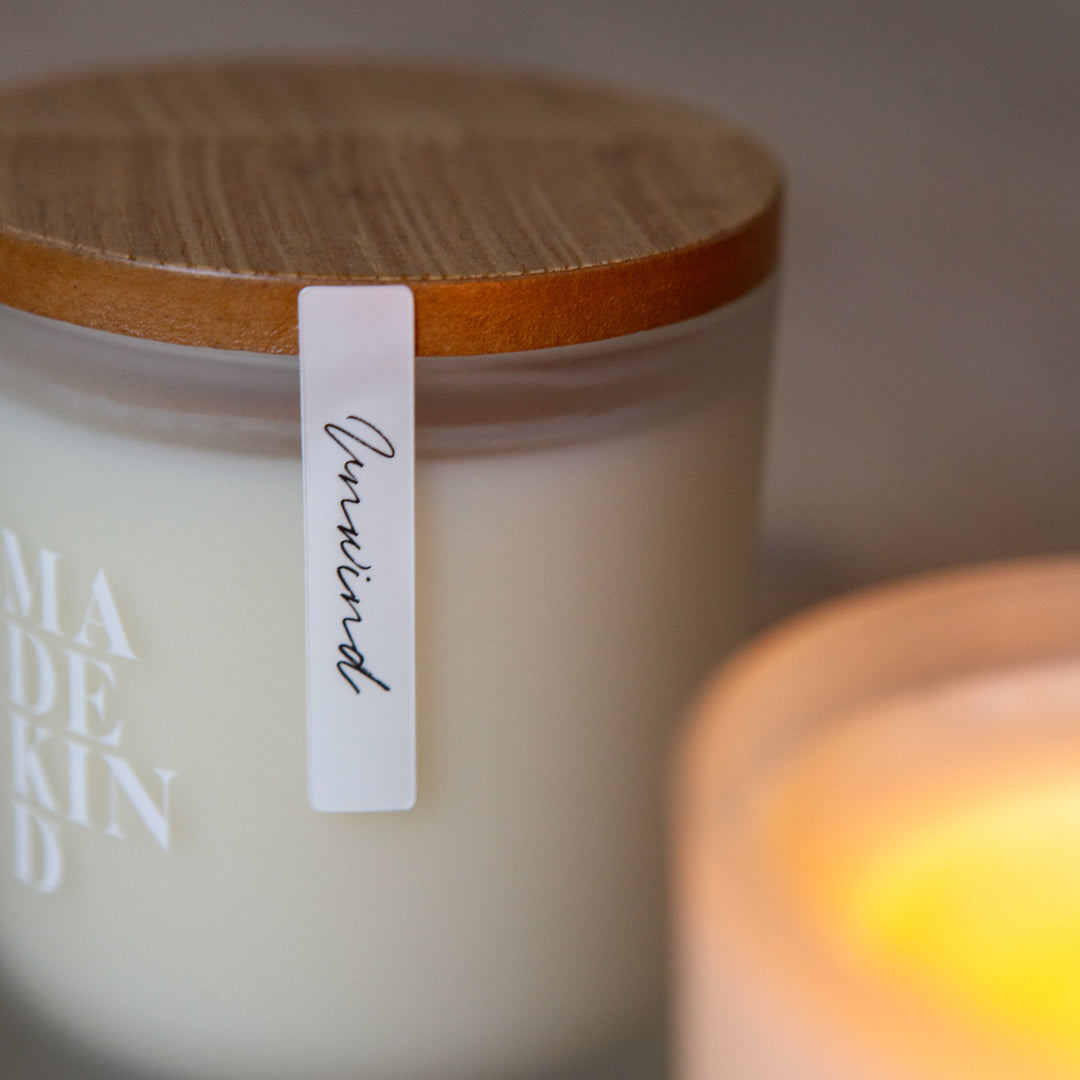 Two candles, one lit and one unlit, on a wooden surface with wooden lid with 'MADEKIND' branding, in unwind blend for relaxation