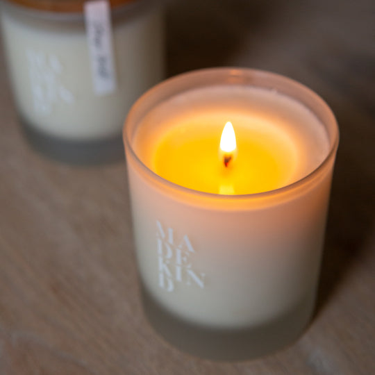 Two candles, one lit and one unlit, on a wooden surface with 'MADEKIND' branding, in unwind blend for relaxation