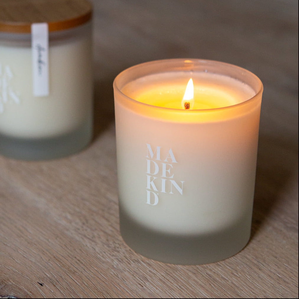 Two candles on a wooden surface, one lit and labeled 'MADE KIND'. Soy wax candle with wood lid.