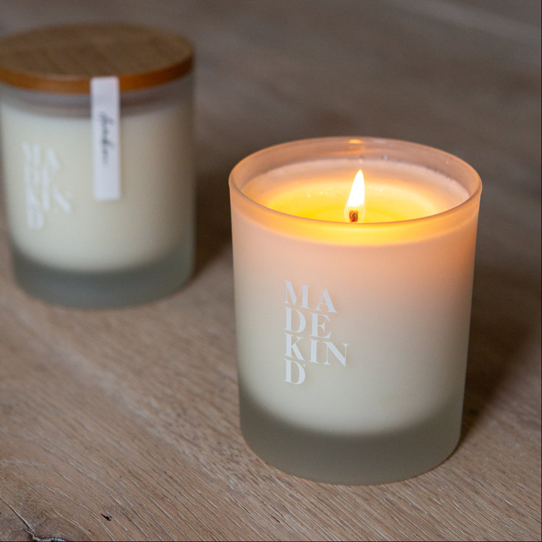 Two candles on a wooden surface, one lit and labeled 'MADE KIND'. Soy wax candle with wood lid.