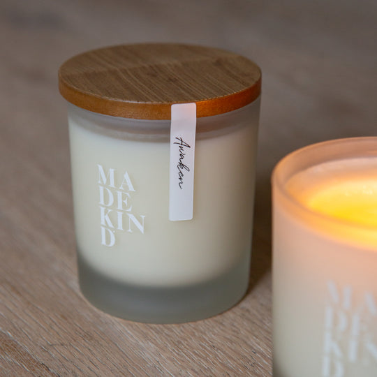 Frosted glass candles on a wooden surface, one lit and labeled 'MADE KIND'. Soy wax candle with wood lid.