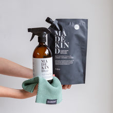 Load image into Gallery viewer, Photo of stretched out arms holding out an amber glass bathroom cleaner with a spray nozzle and a 1 litre refill pouch on a madekind emerald coloured cloth
