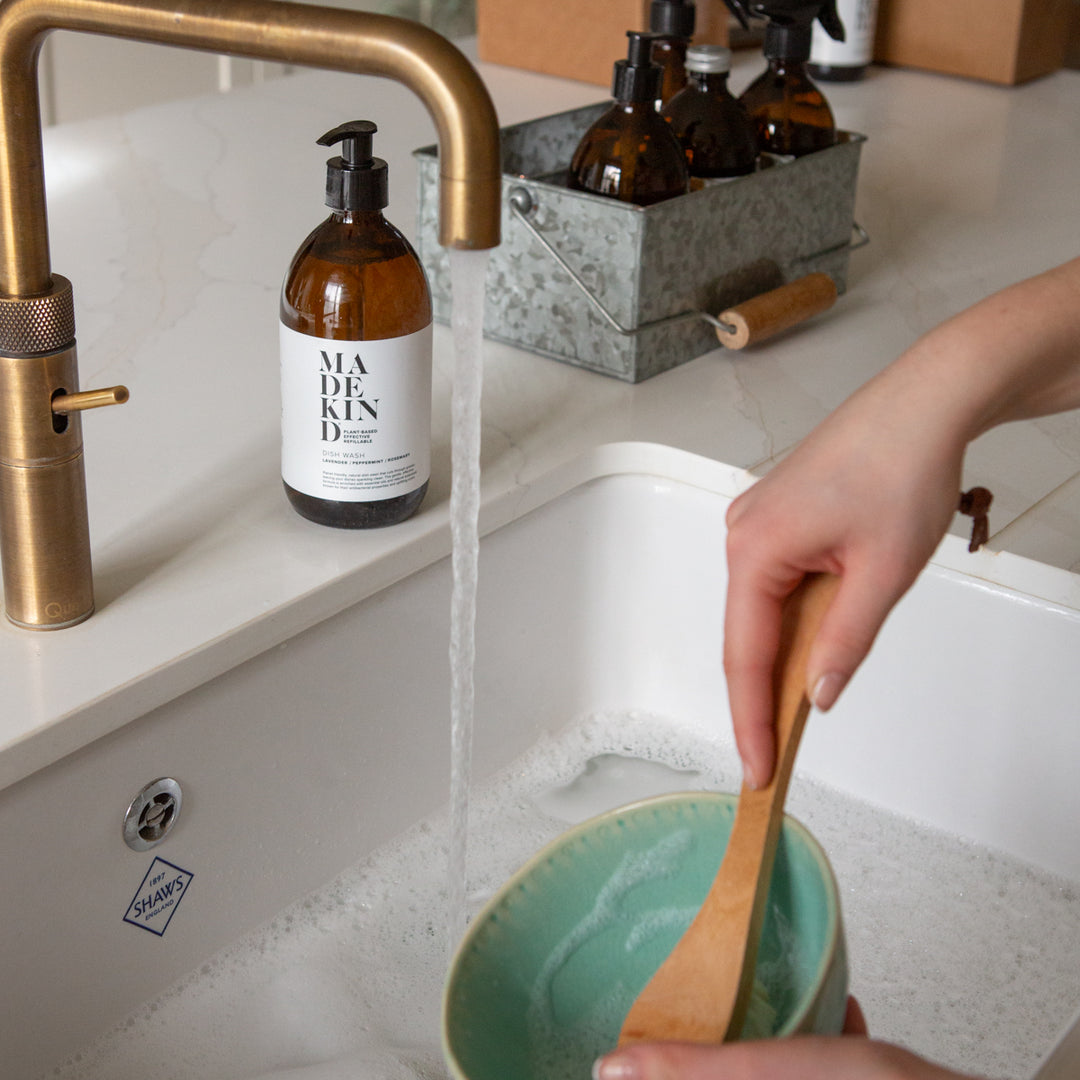 Person washing a green bowl with a wooden spoon in a kitchen sink, with a Madekin bottle and brass faucet in the background.