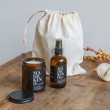 Load image into Gallery viewer, Photo of unwind amber glass candle with the lid of next a room mist spray bottle with a gift bag behind them on a wooden table.
