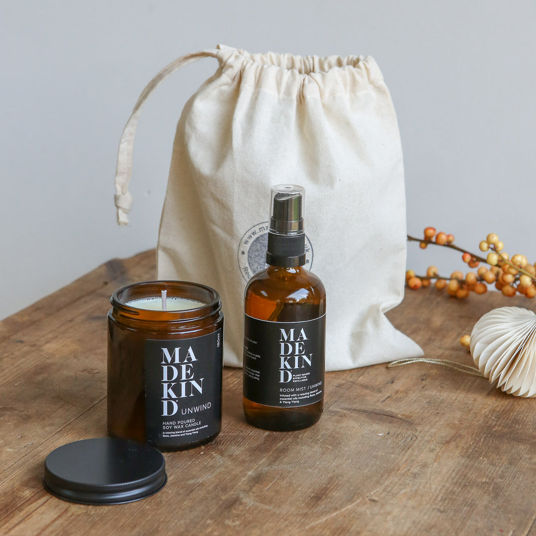 Photo of unwind amber glass candle with the lid of next a room mist spray bottle with a gift bag behind them on a wooden table.